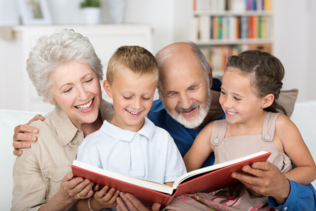 A happily retired couple read a book with their two grandchildren, smiling and enjoying retired life.